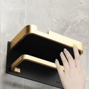 Suporte de papel higiênico preto com dourado, com suporte para telefone e toalhas, fixado na parede do banheiro.
